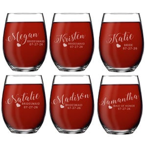 May include: Six stemless wine glasses filled with red liquid. Each glass has a name and title, such as "Megan BRIDESMAID 07-27-26" or "Katie BRIDE 07-27-26", in a decorative font. A heart is also included.