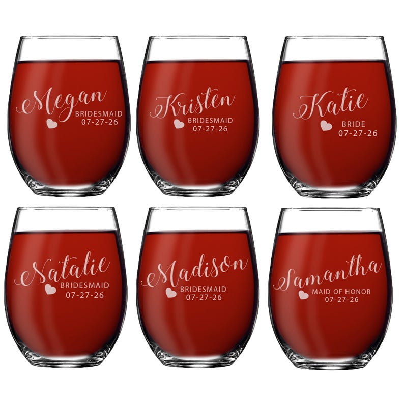 May include: Six stemless wine glasses filled with red liquid. Each glass has a name and title, such as "Megan BRIDESMAID 07-27-26" or "Katie BRIDE 07-27-26", in a decorative font. A heart is also included.