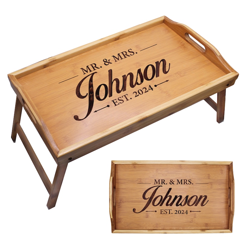 May include: A rectangular bamboo breakfast tray with foldable legs and side handles. The surface features dark laser-engraved text that reads MR. & MRS. Johnson EST. 2024, accented with horizontal lines and arrows. The wood grain is visible throughout.