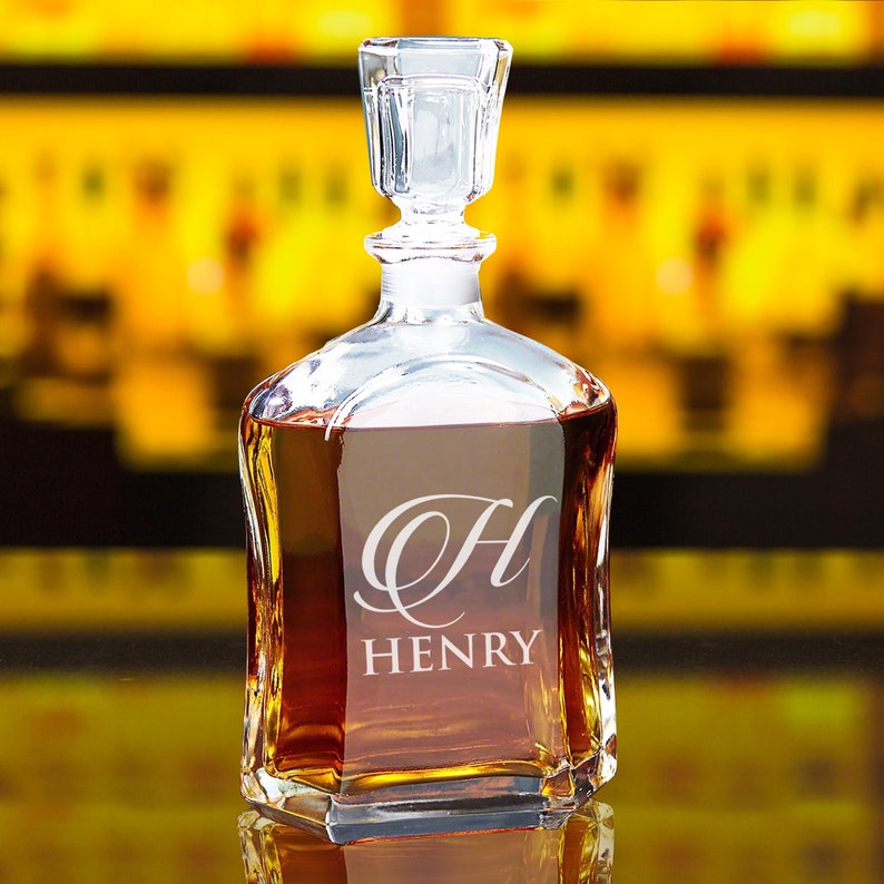 May include: A clear glass decanter with a square base and a matching glass stopper. The front of the decanter features an etched design consisting of a large, stylized letter H above the name HENRY in a clean, capitalized, sans-serif font.