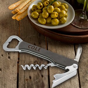 Op de afbeelding: Een multifunctionele flesopener met een kurkentrekker, een flesopener en een klein mes. De tool is gemaakt van metaal en heeft een zwarte handgreep met het woord "COLE" erop gegraveerd.