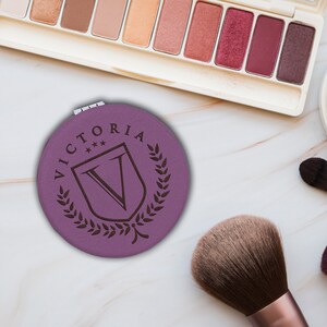 May include: A round purple compact mirror featuring a dark brown engraved design of a shield with the letter V, surrounded by a laurel wreath and the name VICTORIA with three small stars above the shield, including a metal hinge at the top.