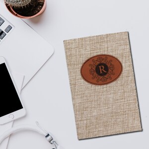 Puede incluir: Un diario beige con textura de lino con un parche de cuero marr&oacute;n. El parche presenta un dise&ntilde;o ornamentado alrededor de la letra "R". El diario est&aacute; sobre una superficie blanca con un port&aacute;til, un tel&eacute;fono y unos auriculares.