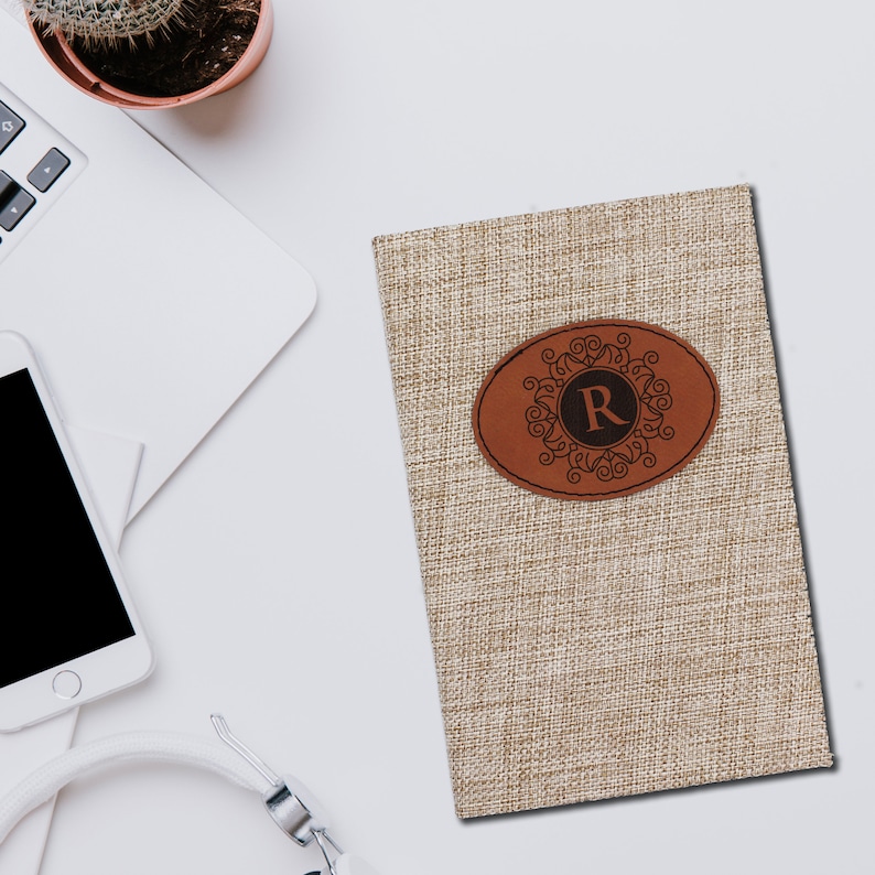 Puede incluir: Un diario beige con textura de lino con un parche de cuero marr&oacute;n. El parche presenta un dise&ntilde;o ornamentado alrededor de la letra "R". El diario est&aacute; sobre una superficie blanca con un port&aacute;til, un tel&eacute;fono y unos auriculares.