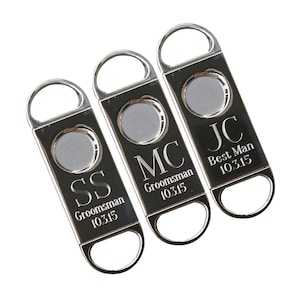 Könnte beinhalten: Drei silberne Zigarrenschneider mit gravierten Initialen und Titeln. Die Schneider sind graviert mit "SS  Groomsman 10315", "MC Groomsman 10315" und "JC Best Man 10315".