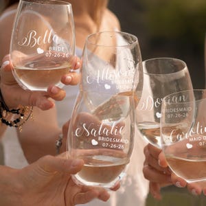 May include: A set of clear stemless wine glasses featuring white printed text and heart icons. Each glass is personalized with names including Bella, Allison, Dorian, Natalie, and Caroline, along with the titles Bride, Bridesmaid, or Maid of Honor and the date 07-26-26.