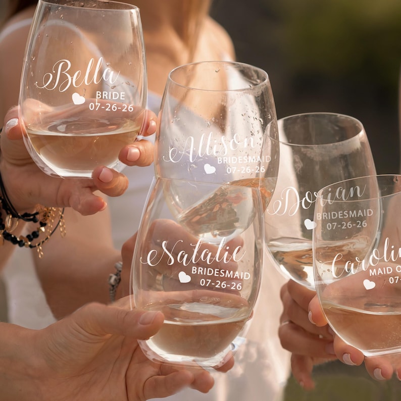May include: A set of clear stemless wine glasses featuring white printed text and heart icons. Each glass is personalized with names including Bella, Allison, Dorian, Natalie, and Caroline, along with the titles Bride, Bridesmaid, or Maid of Honor and the date 07-26-26.