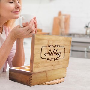K&ouml;nnte beinhalten: Eine rechteckige Teebox aus Bambus mit einem Klappdeckel, der den lasergravierten Namen Ashley in einem dekorativen Rahmen zeigt. Die Box hat sichtbare Fingerzinken an den Ecken und einen offenen Innenraum mit mehreren F&auml;chern.