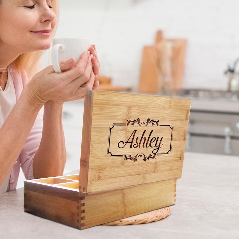 K&ouml;nnte beinhalten: Eine rechteckige Teebox aus Bambus mit einem Klappdeckel, der den lasergravierten Namen Ashley in einem dekorativen Rahmen zeigt. Die Box hat sichtbare Fingerzinken an den Ecken und einen offenen Innenraum mit mehreren F&auml;chern.