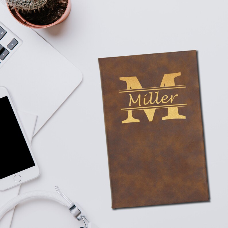 Puede incluir: Un diario de cuero marr&oacute;n con el nombre "Miller" y la letra "M" en letras doradas. El diario es rectangular y parece estar cerrado. Un port&aacute;til, un smartphone y unos auriculares est&aacute;n en el fondo.