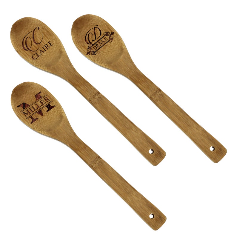 May include: Three bamboo cooking spoons with engraved names and initials. The spoons are light brown with a natural wood grain. One spoon has "Claire" and a "C", another has "Drake" and a "D", and the last has "Miller" and an "M".