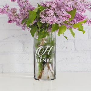 May include: A clear glass vase with the name "Henry" and the letter "H" etched on the front. The vase holds a bouquet of purple lilacs with green leaves. The background is a white brick wall.