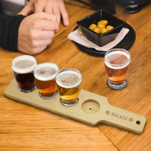 May include: A wooden beer flight paddle with four glasses of beer, each with a different color and foam head. The paddle has a handle with the name "WALKER" engraved on it. A small black bowl of olives sits on the table.