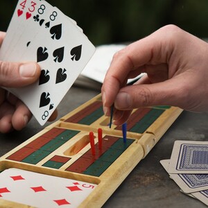 Puede incluir: Un tablero de cribbage de madera con clavijas rojas y azules, una baraja de cartas y una mano de cartas con el 8 de picas, el 3 de tr&eacute;boles y el 4 de corazones.