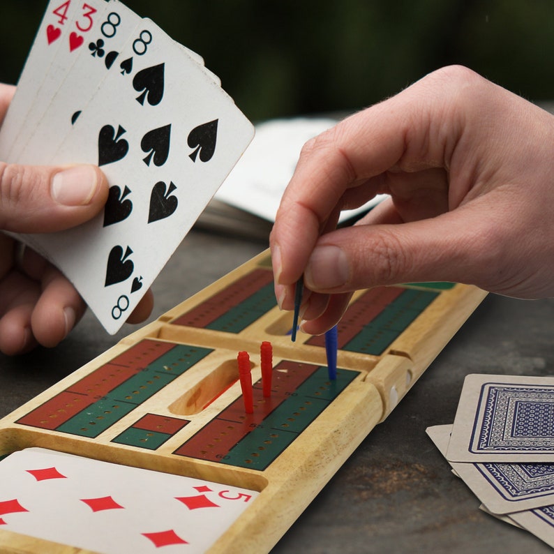 Puede incluir: Un tablero de cribbage de madera con clavijas rojas y azules, una baraja de cartas y una mano de cartas con el 8 de picas, el 3 de tr&eacute;boles y el 4 de corazones.