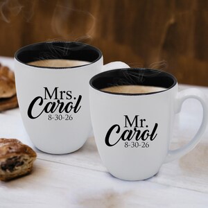 May include: Two white ceramic mugs with black interiors and handles. One mug reads "Mrs. Carol 8-30-26" and the other reads "Mr. Carol 8-30-26" in black script. Both mugs are filled with a hot beverage.