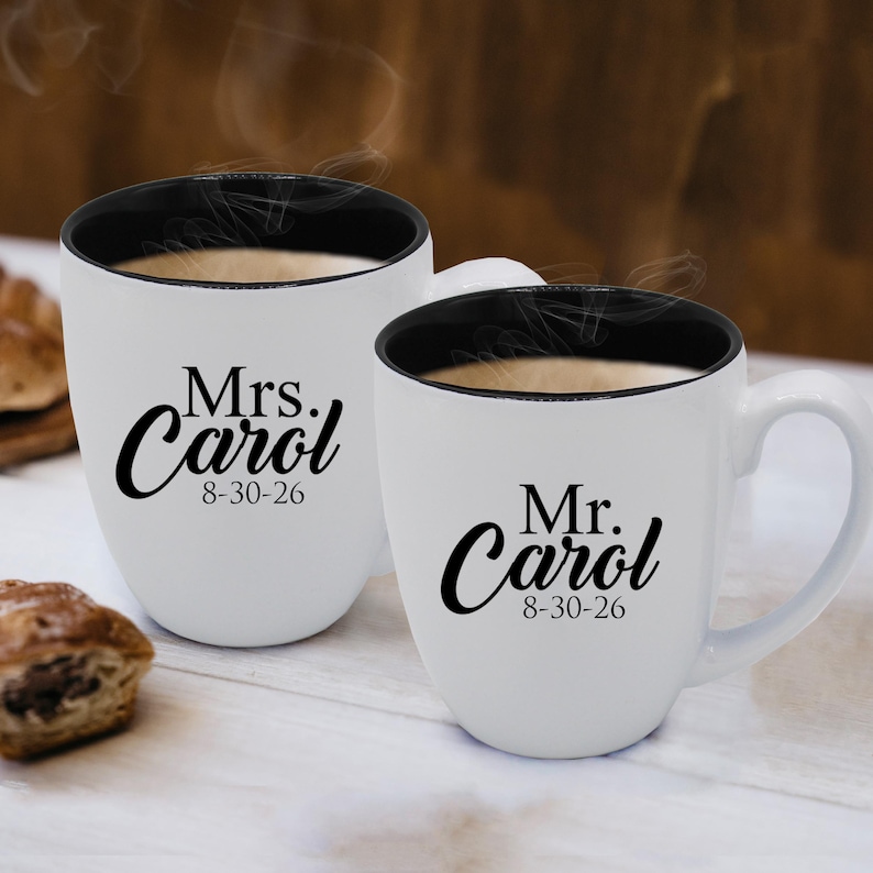 May include: Two white ceramic mugs with black interiors and handles. One mug reads "Mrs. Carol 8-30-26" and the other reads "Mr. Carol 8-30-26" in black script. Both mugs are filled with a hot beverage.