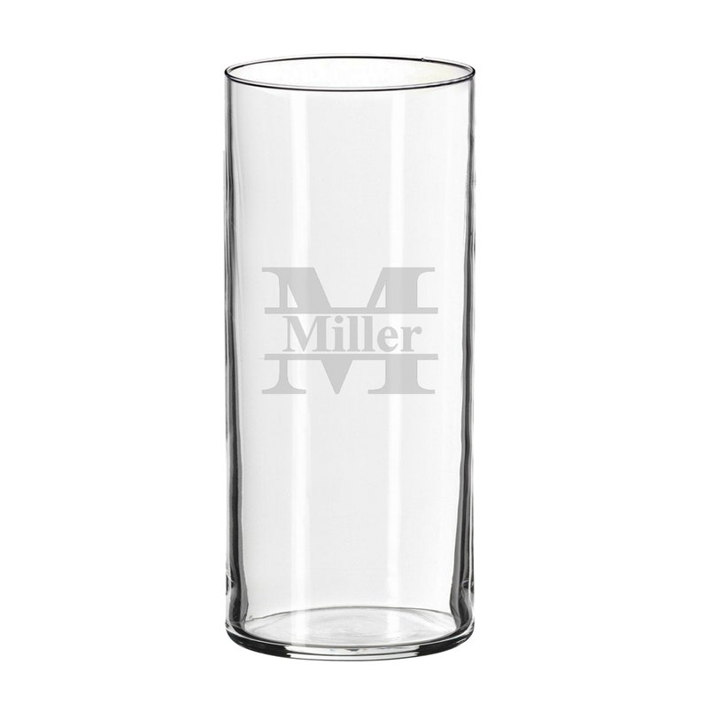 May include: A clear, cylindrical glass with a wide opening. The glass features an etched design of the letter "M" with the name "Miller" below it. The glass is suitable for beverages.