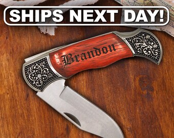 May include: A folding pocket knife with a silver-toned metal blade and a handle featuring a reddish-brown wood grain finish. The handle is engraved with the name Brandon in a black gothic font and has ornate silver metal scrollwork at both ends.