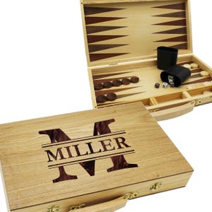 May include: A wooden backgammon set with a personalized design. The lid features the name "MILLER" in a bold, dark brown font, with a large initial "M". The set is open, revealing the game board, dice, and playing pieces. The set is made of light-colored wood.