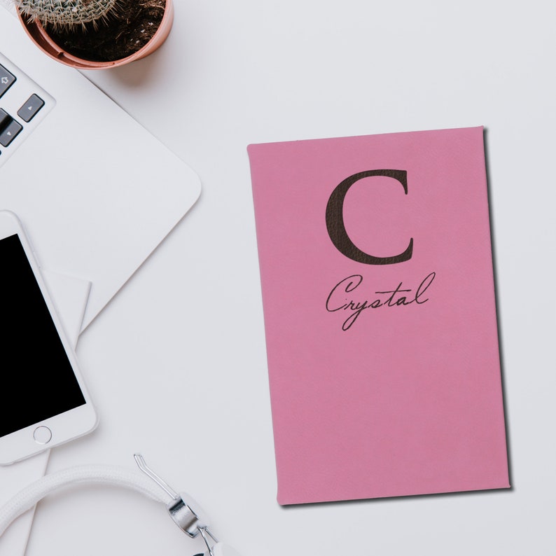 Puede incluir: Un cuaderno rosa con la letra "C" y el nombre "Crystal" impresos en la portada. El cuaderno est&aacute; sobre una superficie blanca, junto a un port&aacute;til, un smartphone y unos auriculares. Tambi&eacute;n se ve un peque&ntilde;o cactus en maceta.