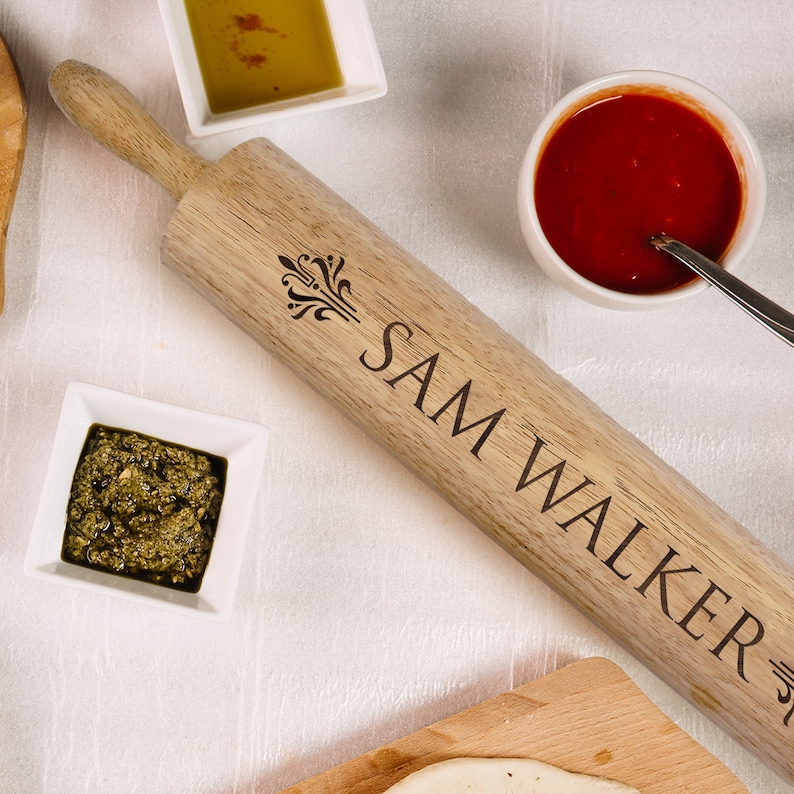 Puede incluir: Un rodillo de madera con el nombre "SAM WALKER" grabado y un dise&ntilde;o decorativo. La imagen incluye peque&ntilde;os cuencos blancos llenos de pesto, aceite de oliva y salsa de tomate, lo que sugiere un tema culinario.