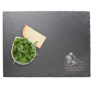 May include: A rectangular dark gray slate serving board with rough, uneven edges. The board features an engraved design of a grape cluster with vines and the text "SIMPSON EST. 2019". A white bowl filled with green arugula and a wedge of cheese are present.