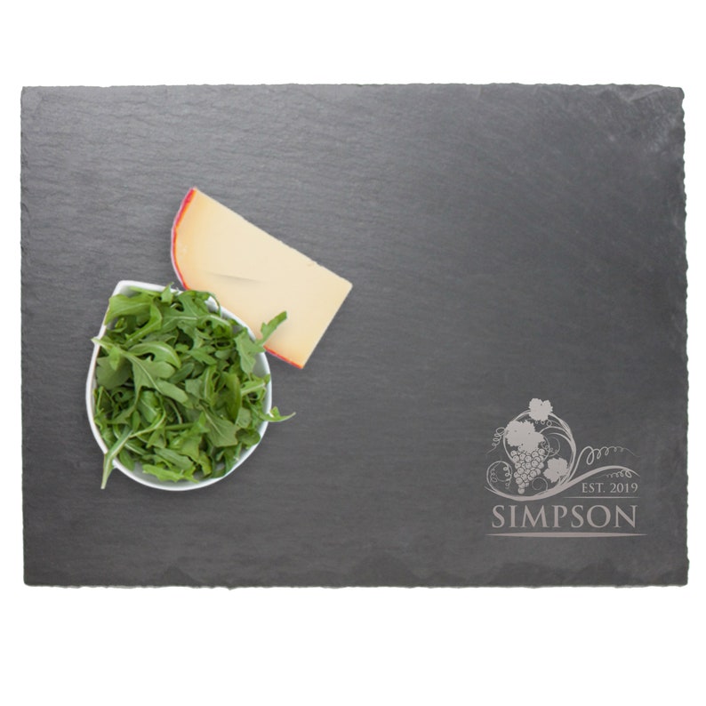 May include: A rectangular dark gray slate serving board with rough, uneven edges. The board features an engraved design of a grape cluster with vines and the text "SIMPSON EST. 2019". A white bowl filled with green arugula and a wedge of cheese are present.