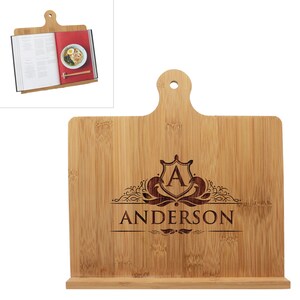 May include: A light brown bamboo cookbook stand featuring a handle with a circular cutout at the top and a horizontal ledge at the base. The center is laser-engraved with a decorative crest containing the letter A and the name ANDERSON below.