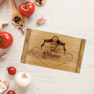 Pu&ograve; includere: Un tagliere rettangolare in bamb&ugrave; con un design inciso al laser che presenta l'icona di un cappello da chef, la lettera W in un cerchio e il testo "SINCE 2016", "Warner" e "FAMILY RECIPES" centrati sulla superficie in legno chiaro.