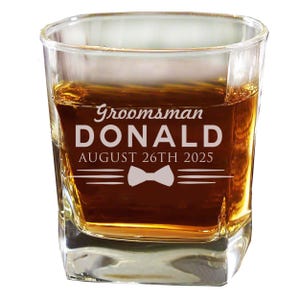 May include: A clear glass rocks glass with a personalized inscription that reads "Groomsman DONALD AUGUST 26TH 2025" with a bow tie graphic.