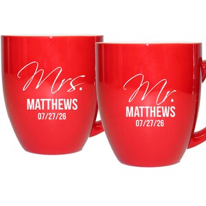 May include: Two red ceramic mugs with white text. One mug reads "Mrs. MATTHEWS 07/27/26" and the other reads "Mr. MATTHEWS 07/27/26". Both mugs have a curved shape and a handle.