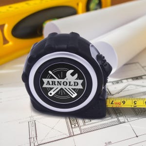 May include: A black and silver tape measure with the name "ARNOLD" and a wrench and screwdriver logo. The tape measure is on top of a blueprint with a yellow level and rolled-up white paper in the background. The tape measure shows measurements in inches.