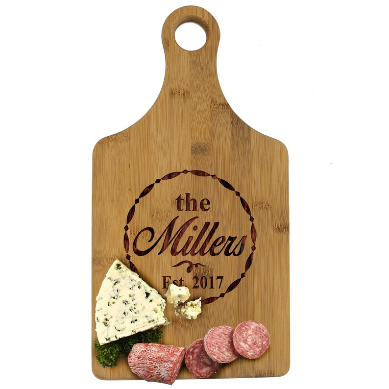 May include: A personalized bamboo cutting board with a round engraved design that says "the Millers Est. 2017". The board has a handle and is shown with cheese and salami on top.