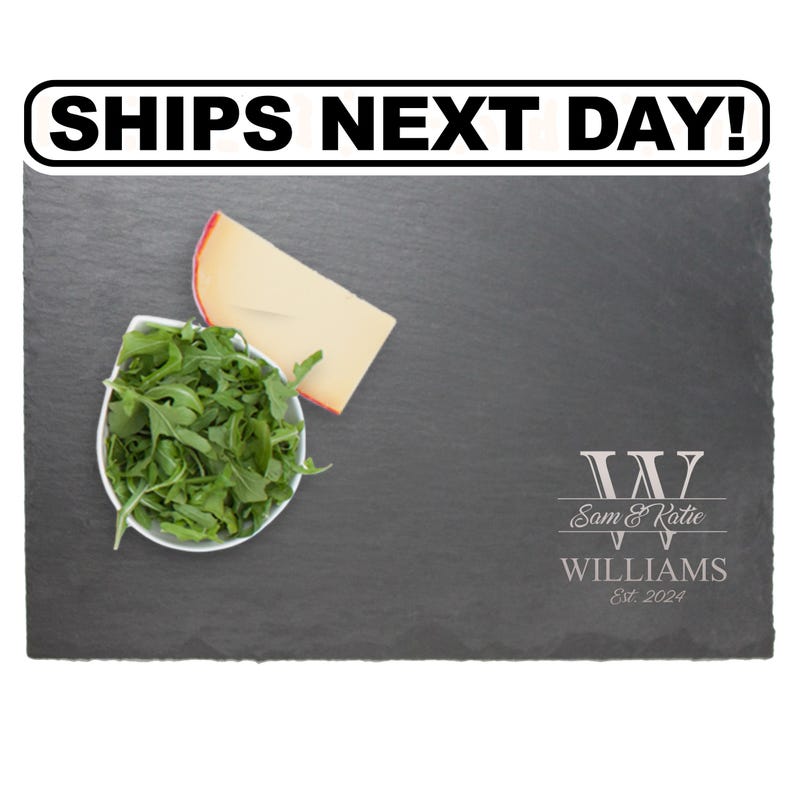 May include: A rectangular black slate serving board featuring a white bowl of green arugula and a wedge of yellow cheese. The board is engraved with a large letter W, the names Sam & Katie, the surname WILLIAMS, and the text Est. 2024.