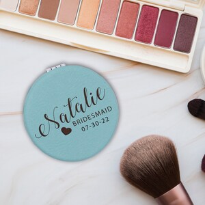 May include: A round, light blue compact mirror featuring the name Natalie in dark brown script, with the text BRIDESMAID and the date 07-30-22 printed below, accompanied by a small dark brown heart icon on the surface of the mirror casing.