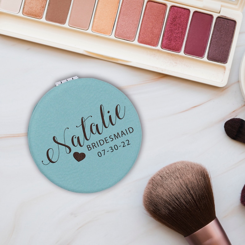 May include: A round, light blue compact mirror featuring the name Natalie in dark brown script, with the text BRIDESMAID and the date 07-30-22 printed below, accompanied by a small dark brown heart icon on the surface of the mirror casing.