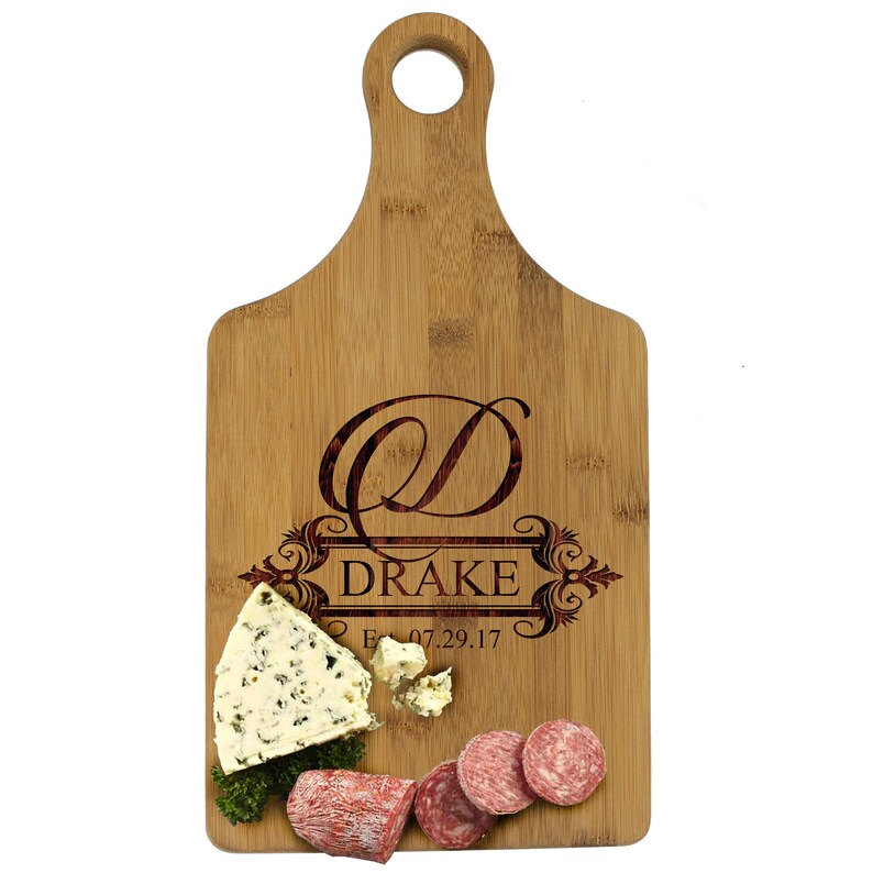 May include: Personalized bamboo cutting board with a monogrammed "D" and the name "Drake" engraved in a decorative font. The board also includes the date "Est. 07.29.17". The board is shown with cheese and salami on top.