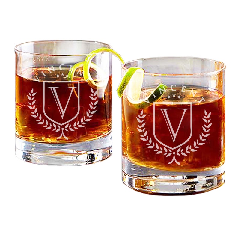 May include: Two clear glass whiskey tumblers with a personalized monogram design. Each glass features a shield with the letter "V" inside, surrounded by a laurel wreath. The text "Vincent" is etched above the shield.