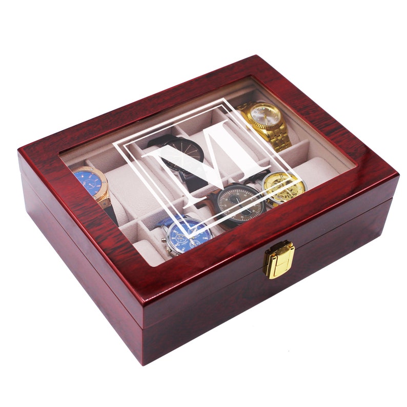 May include: A rectangular dark cherry wood watch box with a glass lid featuring a white letter M inside a square border. The interior is lined with beige fabric and contains multiple compartments holding various wristwatches with metal bands.