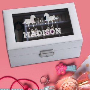 May include: A white rectangular jewellery box with a glass lid featuring a silver graphic of two horses, a horseshoe, and the name MADISON. The box has a silver metal latch and internal compartments visible through the glass top.