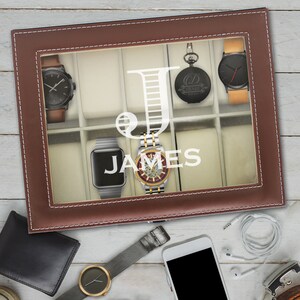May include: A brown leather watch display case with a glass lid featuring a white letter J and the name JAMES. The case contains several wristwatches and a pocket watch. Outside the case are a wallet, a smartphone, earbuds, and a watch.