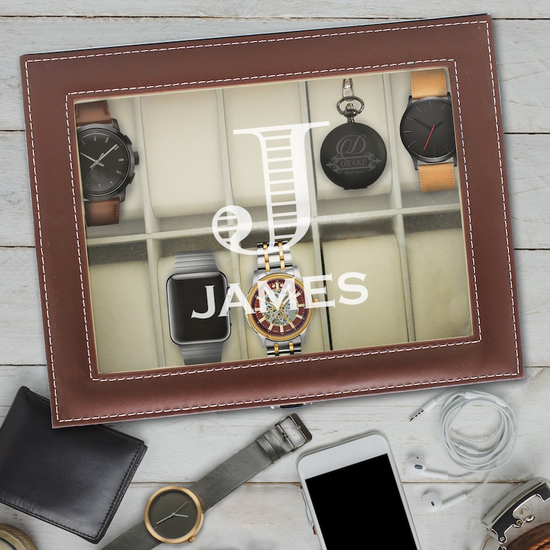 May include: A brown leather watch display case with a glass lid featuring a white letter J and the name JAMES. The case contains several wristwatches and a pocket watch. Outside the case are a wallet, a smartphone, earbuds, and a watch.