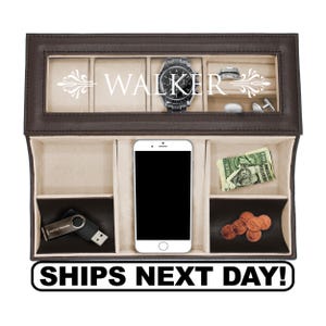 May include: A dark brown leather valet tray with a glass lid featuring the white text WALKER. The interior has beige velvet compartments containing a wristwatch, ring, cufflinks, a smartphone, a USB drive, paper currency, and copper coins.