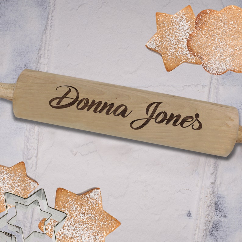 Puede incluir: Un rodillo de madera de color claro con el nombre "Donna Jones" grabado est&aacute; centrado en la imagen. Galletas en forma de estrella y flor espolvoreadas con az&uacute;car glas est&aacute;n dispersas alrededor del rodillo, junto con cortadores de galletas en forma de estrella. Tema de reposter&iacute;a.