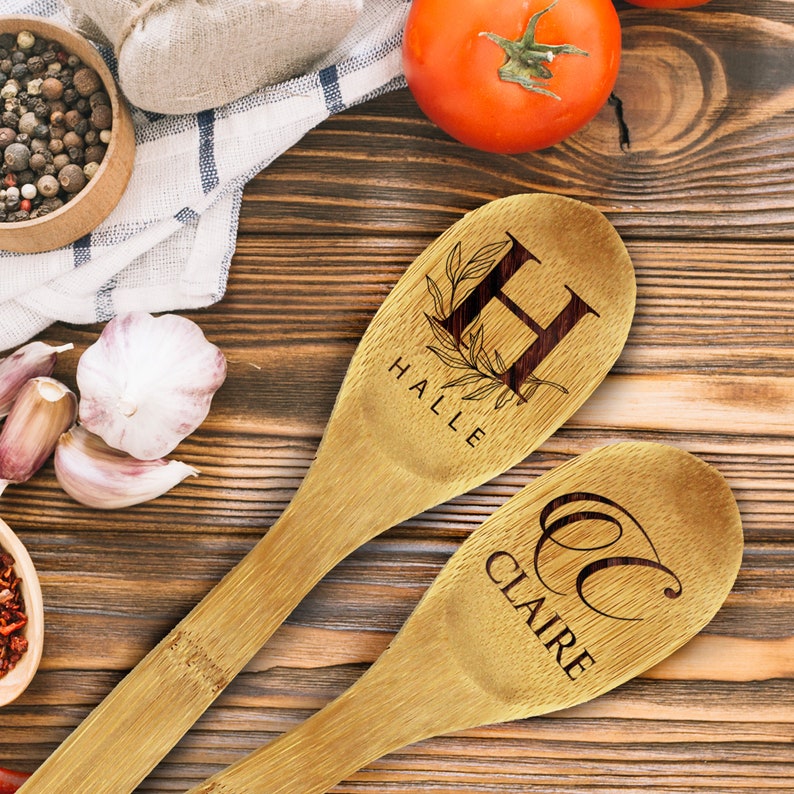 May include: Two wooden cooking spoons with engraved lettering. One spoon features the letter "H" with the name "HALLE" below, and the other has the letter "C" with the name "CLAIRE". The spoons are light brown and rest on a wooden surface.