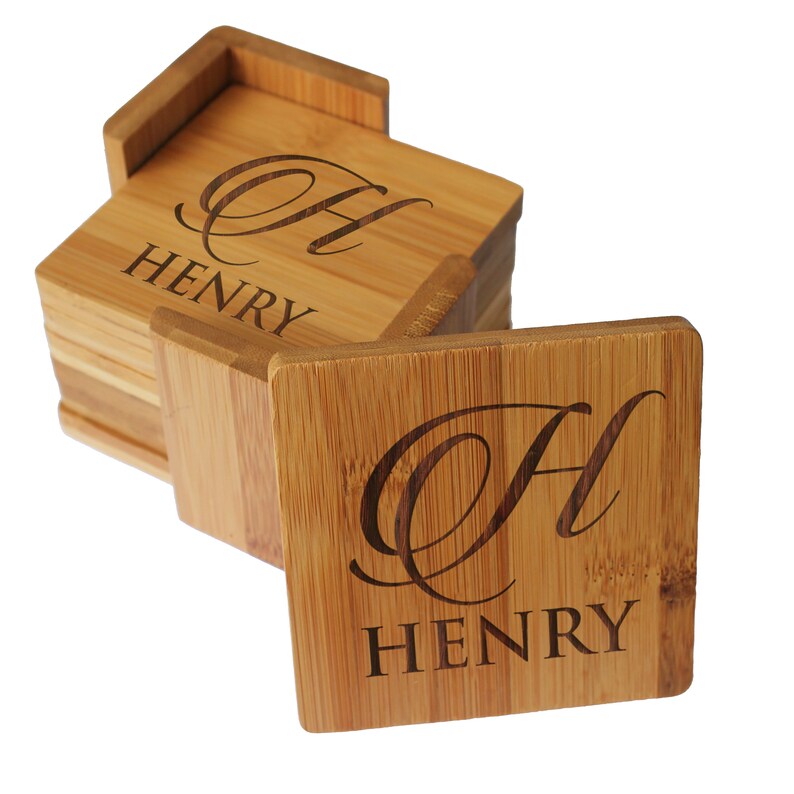 May include: A stack of square bamboo coasters with a matching holder. Each coaster features a laser-engraved design consisting of a large cursive letter H positioned above the name HENRY in a bold, capitalized, sans-serif typeface.