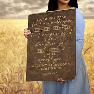 Puede incluir: Un letrero rectangular de madera marrón con el texto "SO DO NOT FEAR for I am with you" y otras palabras inspiradoras de Isaías 41:10. El letrero lo sostiene una persona con un vestido azul, con un campo de trigo al fondo.