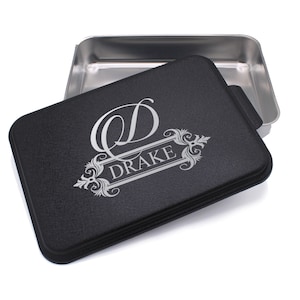 May include: A black baking dish with a silver lid. The lid has a silver monogram with the letter "D" and the name "DRAKE" engraved on it.