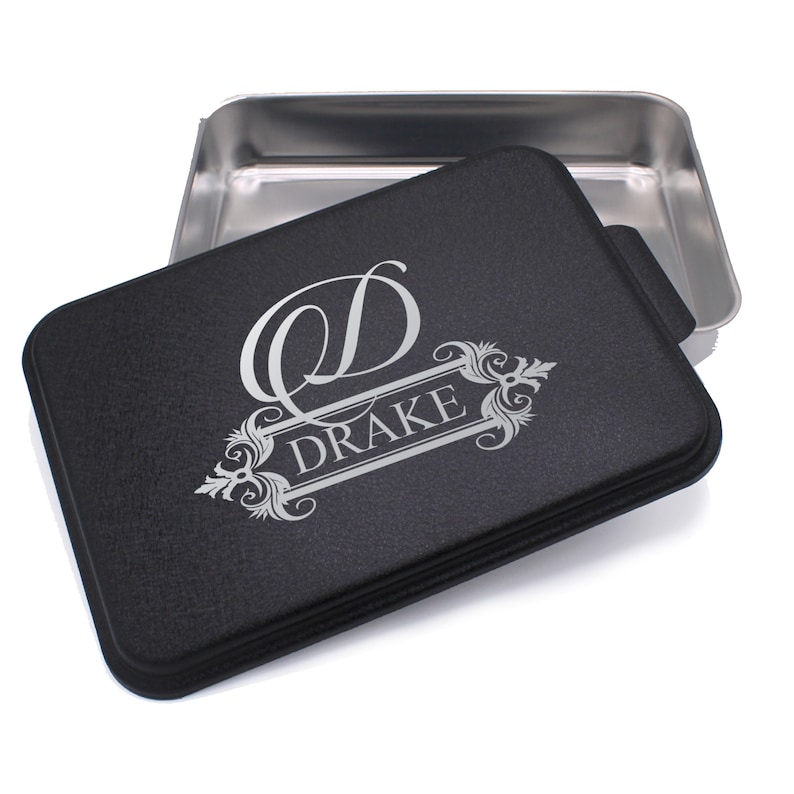 May include: A black baking dish with a silver lid. The lid has a silver monogram with the letter "D" and the name "DRAKE" engraved on it.